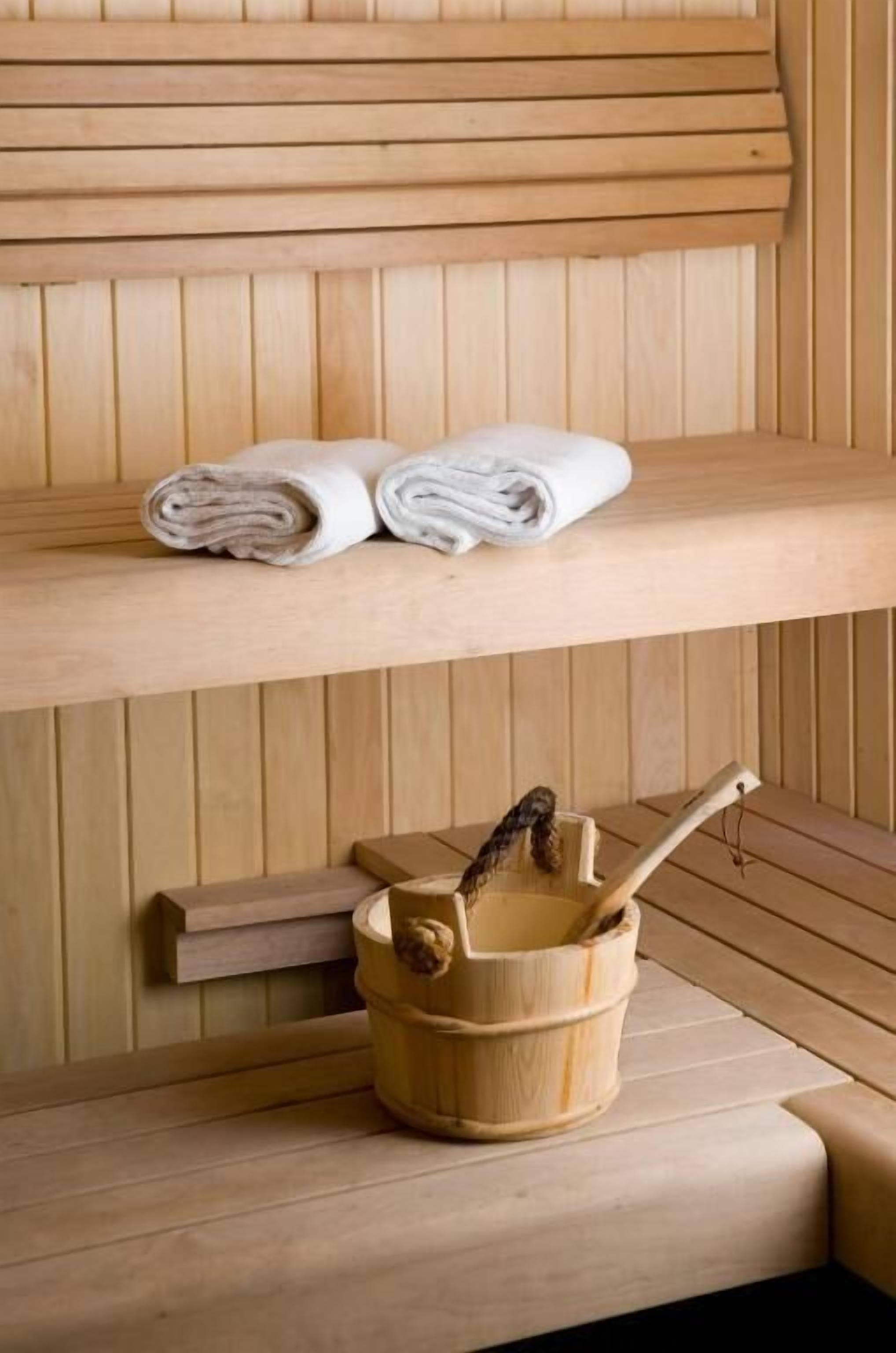 A wooden bench with a shelf above it holding rolled towels and a wooden bucket with a brush.