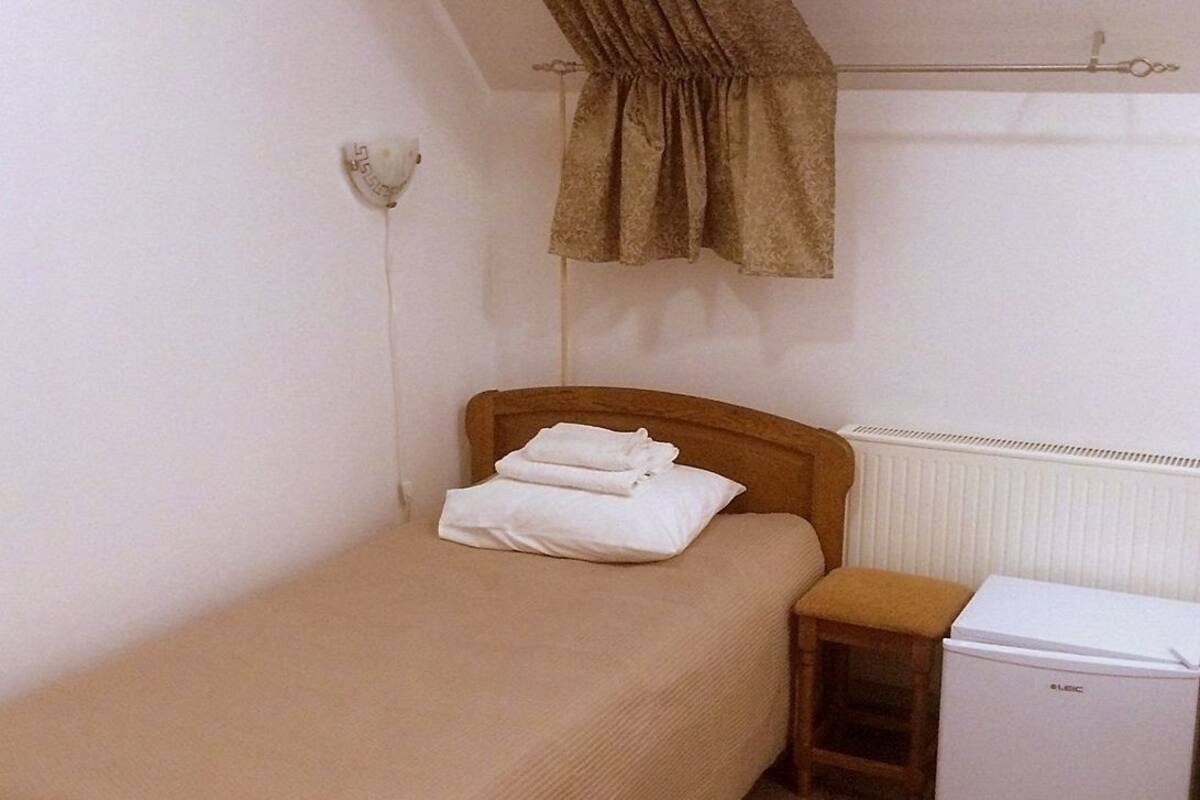 A small, single-bed room with a wooden headboard, a small bedside table, a white radiator, and a white mini-fridge.