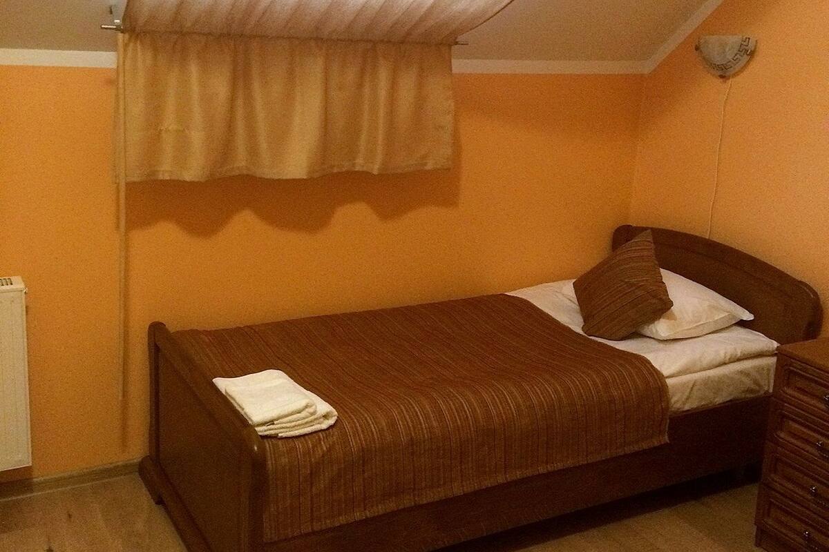 A single bed with a brown headboard and a white blanket, a wooden nightstand, and a beige curtain.