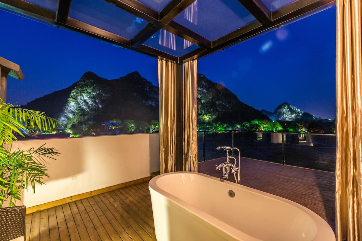 A modern bathroom with a freestanding bathtub, wooden flooring, and a view of mountains at night.