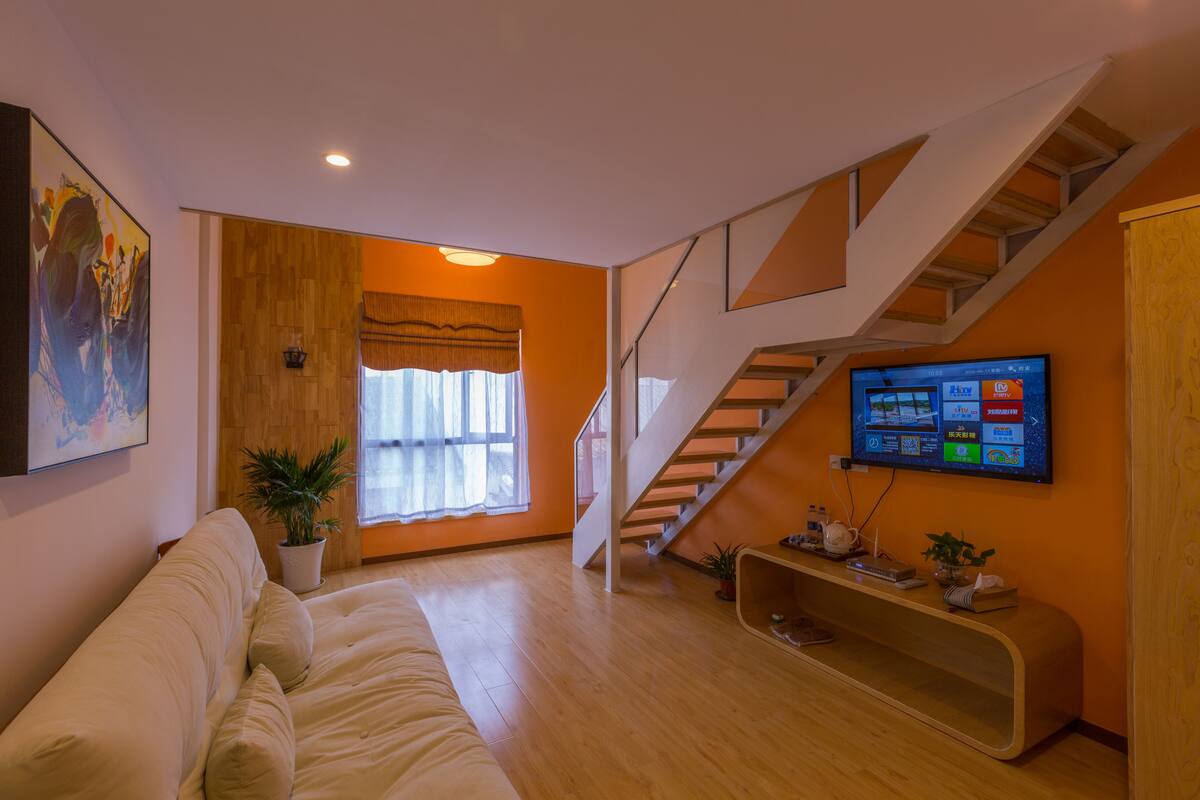 A modern living room with a wooden floor, a sofa, a staircase, and a flat-screen TV mounted on the wall.