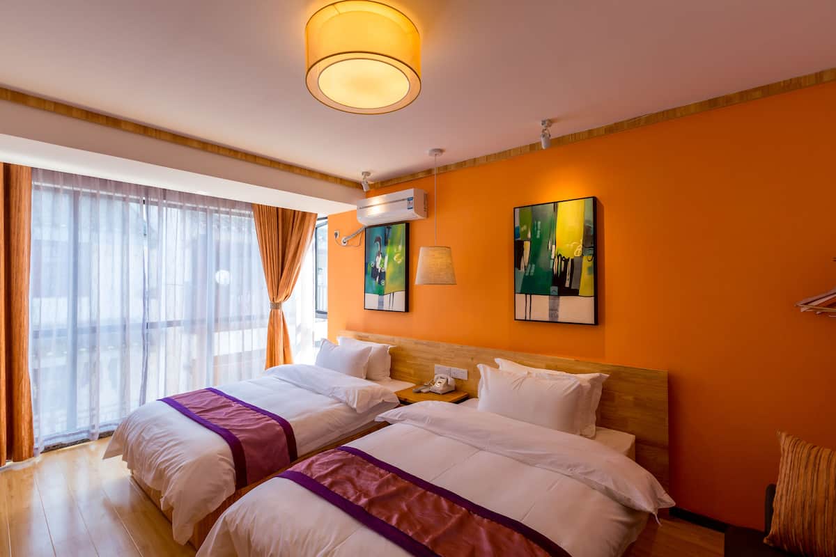 A hotel room with two beds, a large window with sheer curtains, an air conditioning unit, and artwork on the walls.