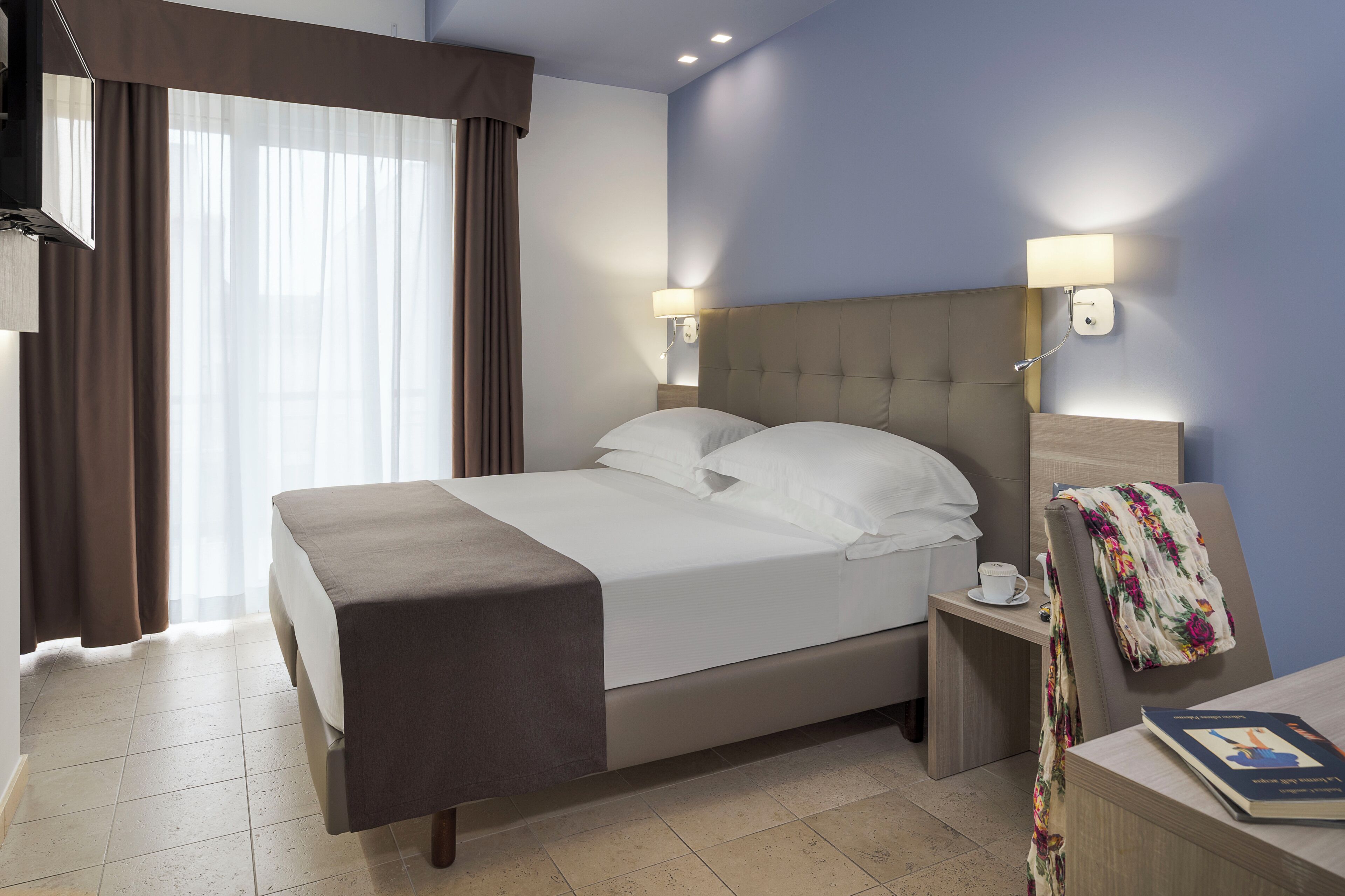 Ein Hotelzimmer mit einem Bett, zwei Nachttischen, einem Sessel mit einer floralen Decke und einem Buch auf dem Nachttisch.