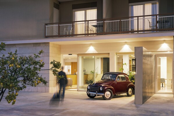 A building entrance with a classic car parked outside, a balcony with a railing, and a reception desk. - Netum Hotel (Noto)