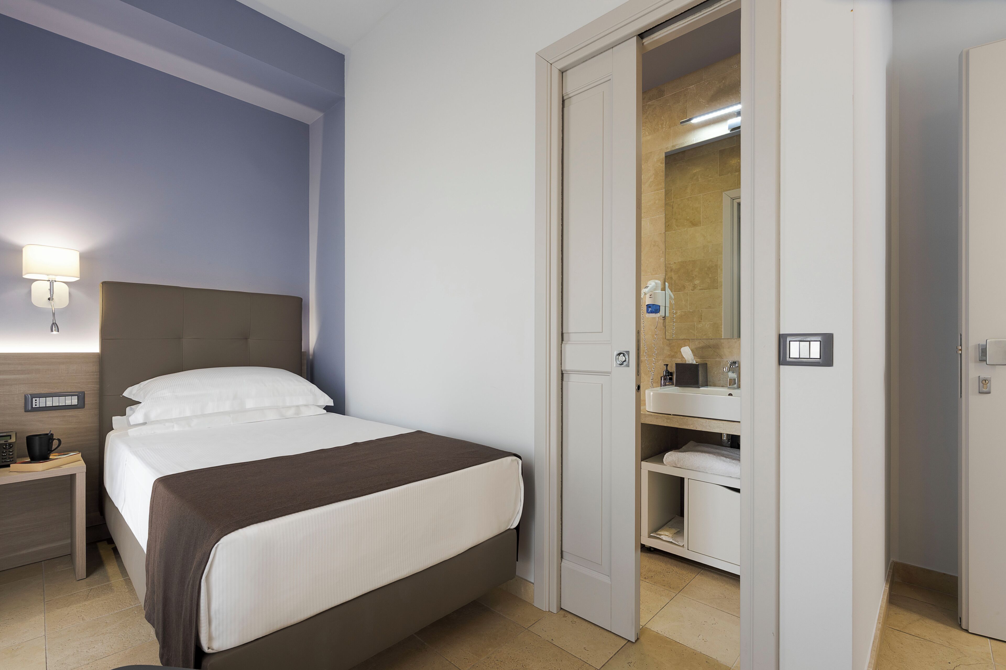 Une chambre d’hôtel avec un lit, une table de chevet, une lampe et une salle de bain visible à travers une porte ouverte.