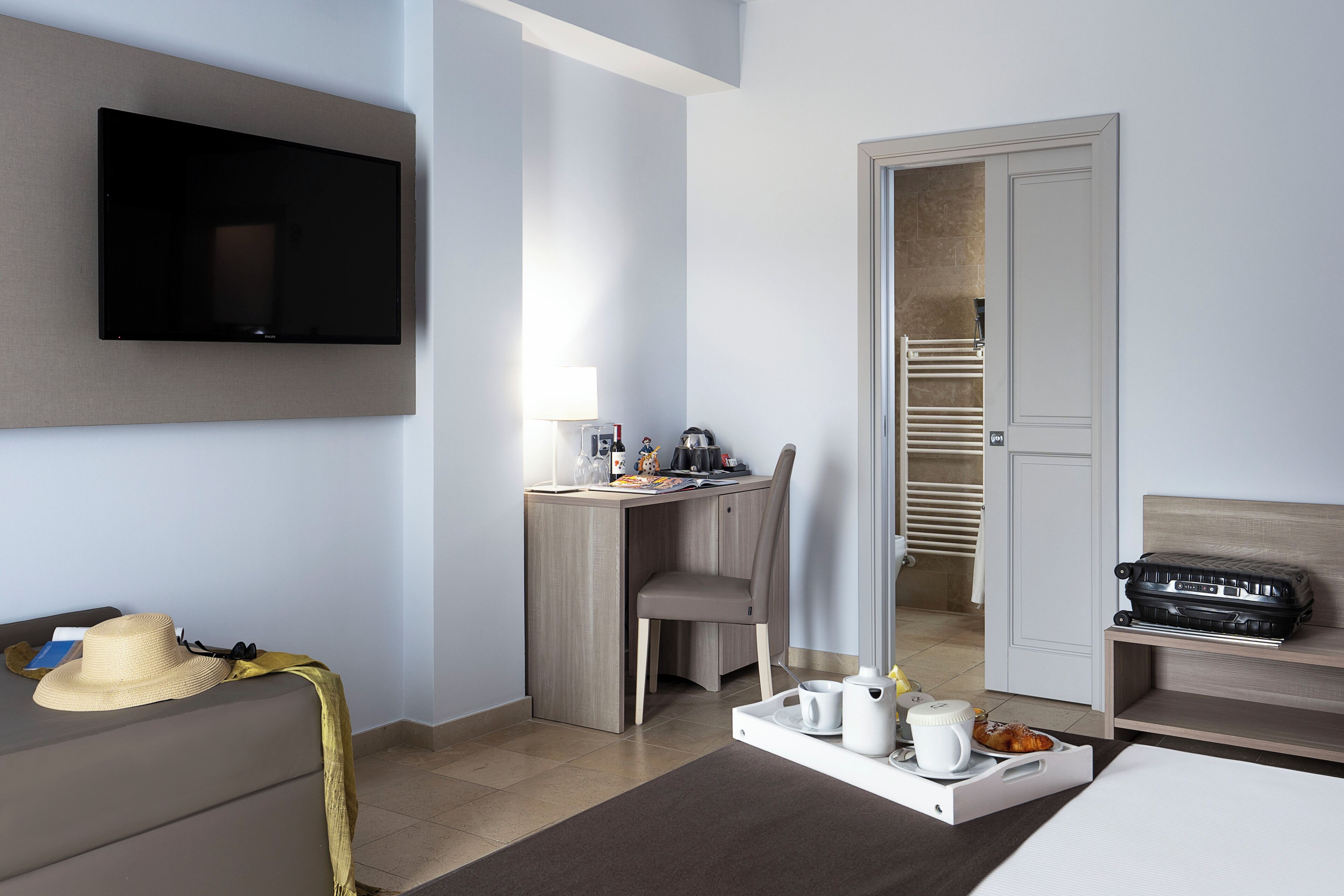 A hotel room with a flat-screen TV, a desk with a chair, a bed, a suitcase, and a tray with breakfast items.