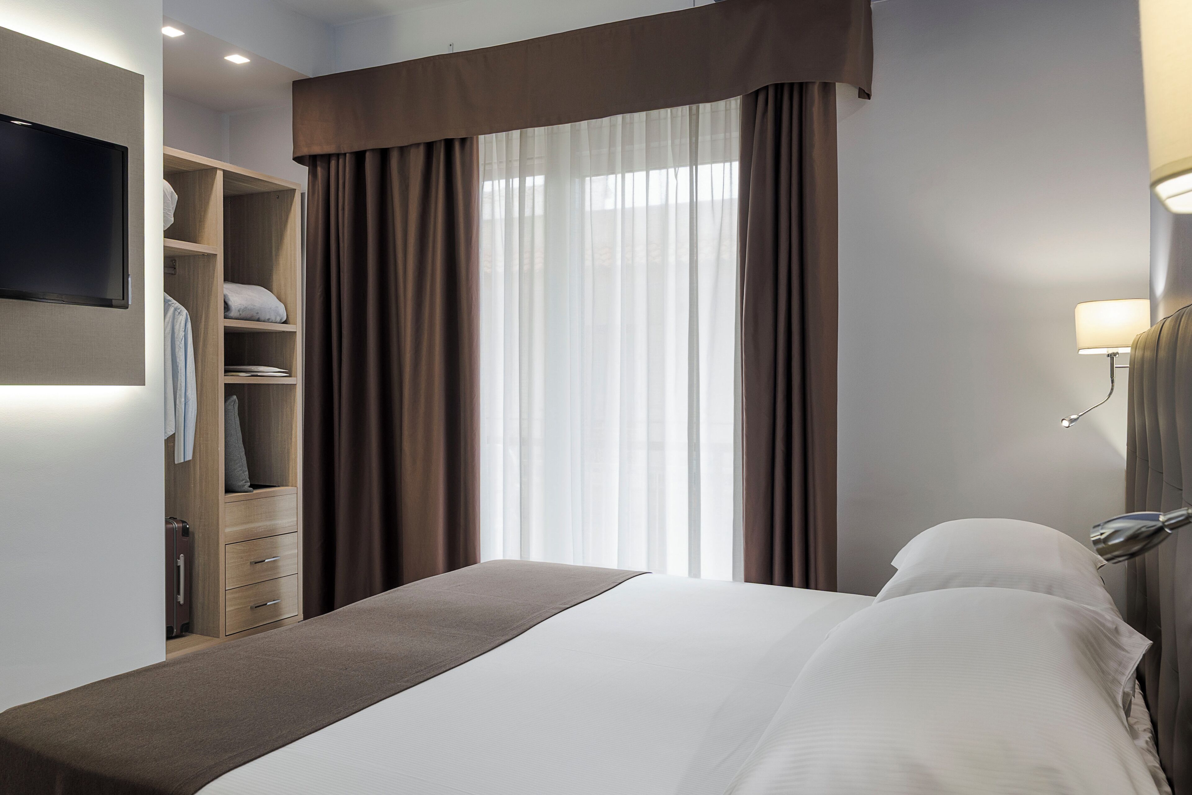 Une chambre d’hôtel avec un lit, un téléviseur fixé au mur, une armoire et une fenêtre avec des rideaux.
