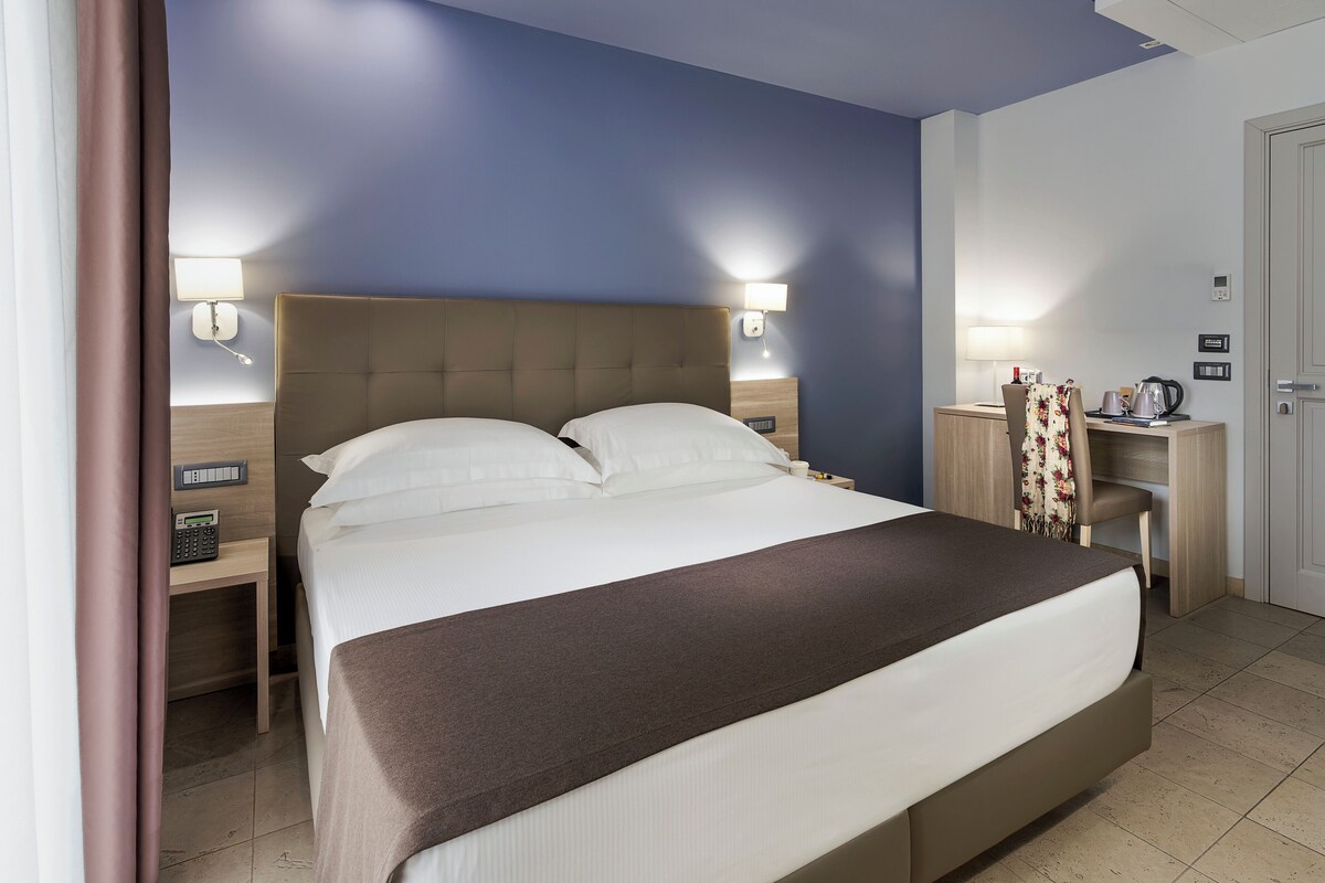 A hotel room with a large bed, bedside tables, a desk, and a wardrobe.