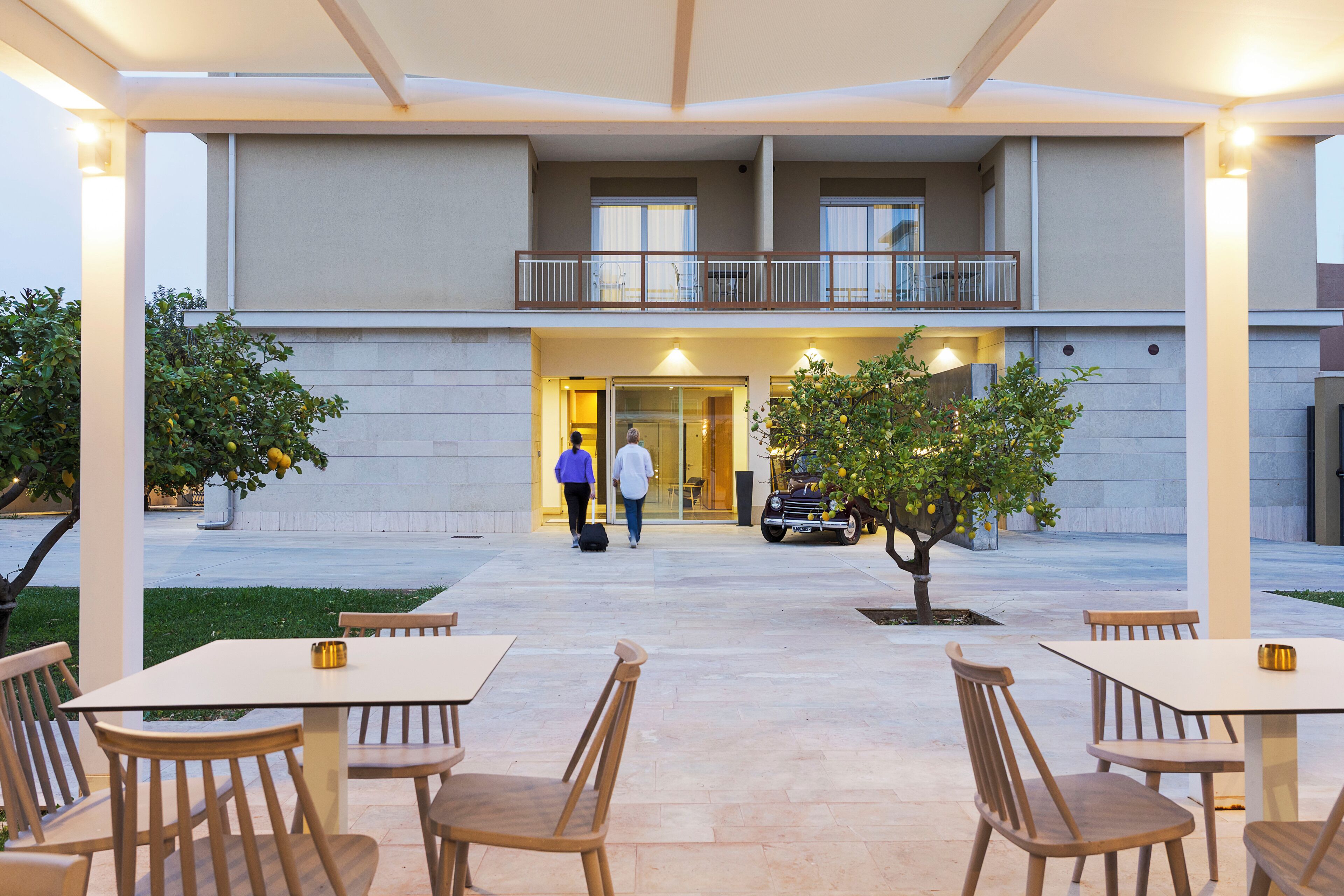 Entrada de hotel con un área de estar al aire libre techada, un edificio de dos pisos y un árbol en primer plano.