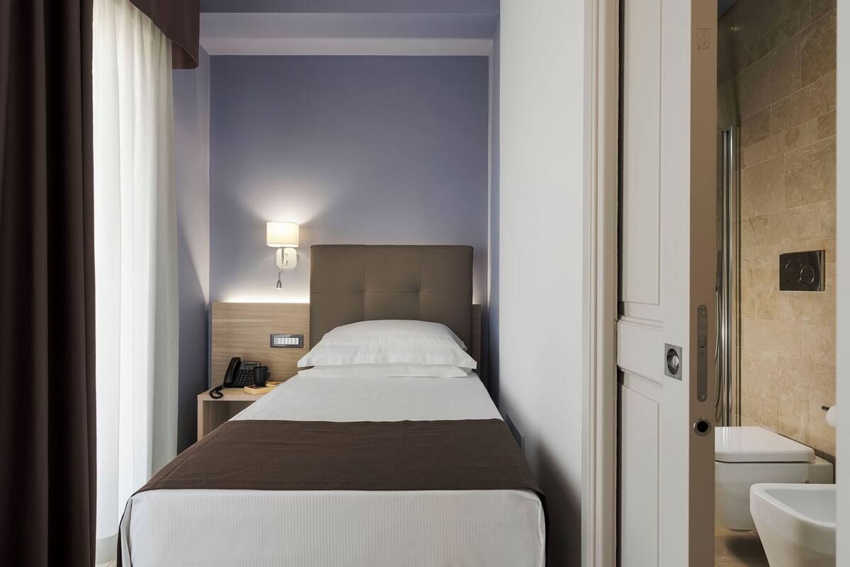 A hotel room with a bed, bedside table with a lamp, and a bathroom with a toilet and sink.