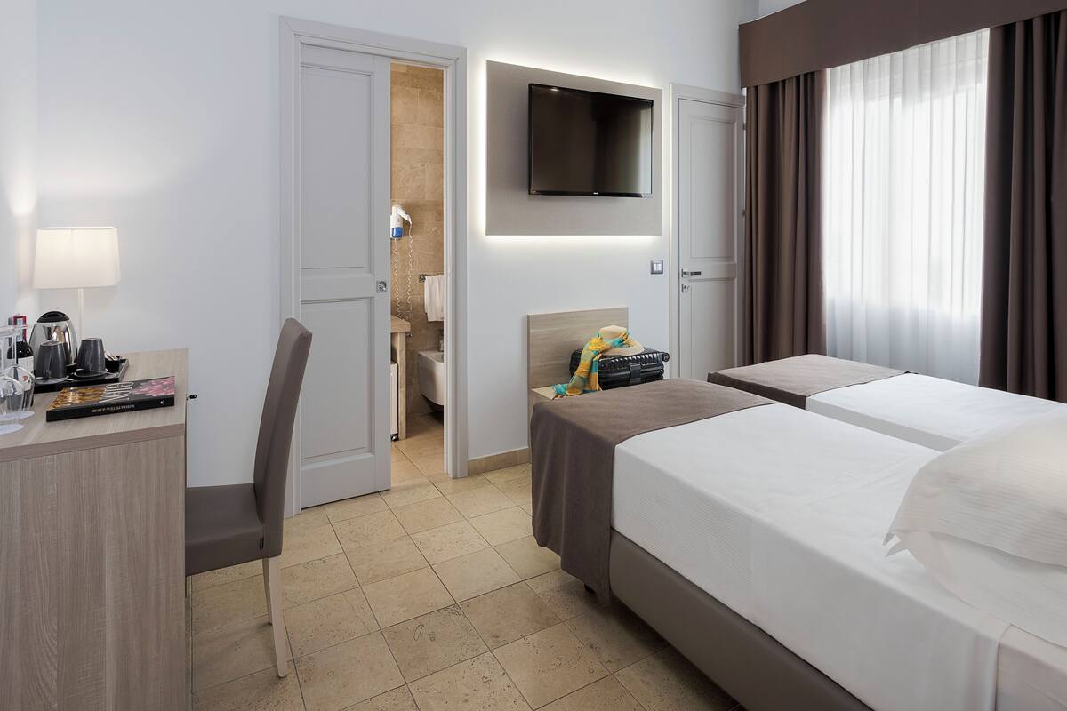 A hotel room with a bed, a desk, a chair, a TV mounted on the wall, and a bathroom visible through an open door.