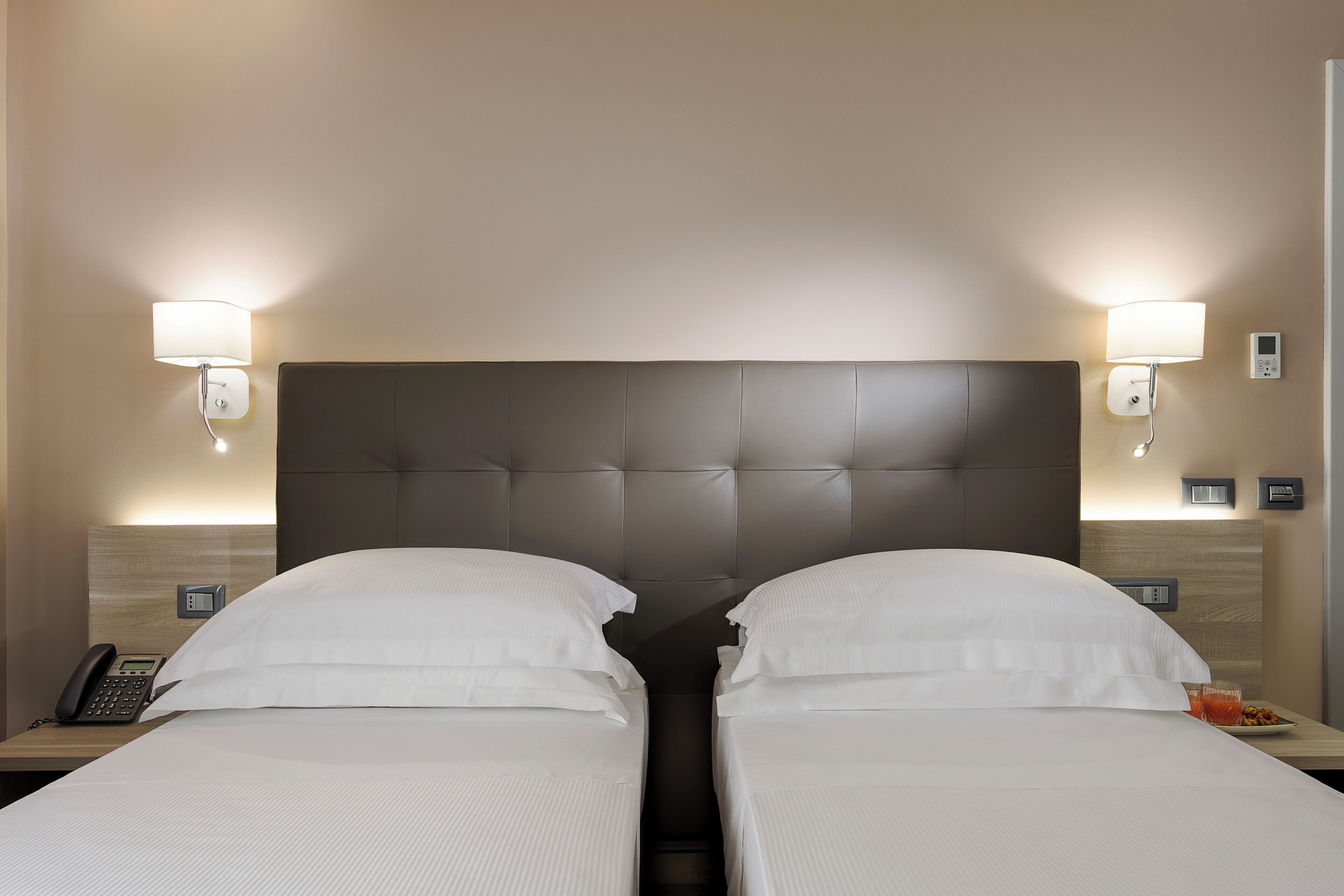 Ein Hotelzimmer mit zwei Betten, einem gepolsterten Kopfteil, Nachttischlampen, einem Telefon und einem kleinen Tisch mit einem Glas Wasser.