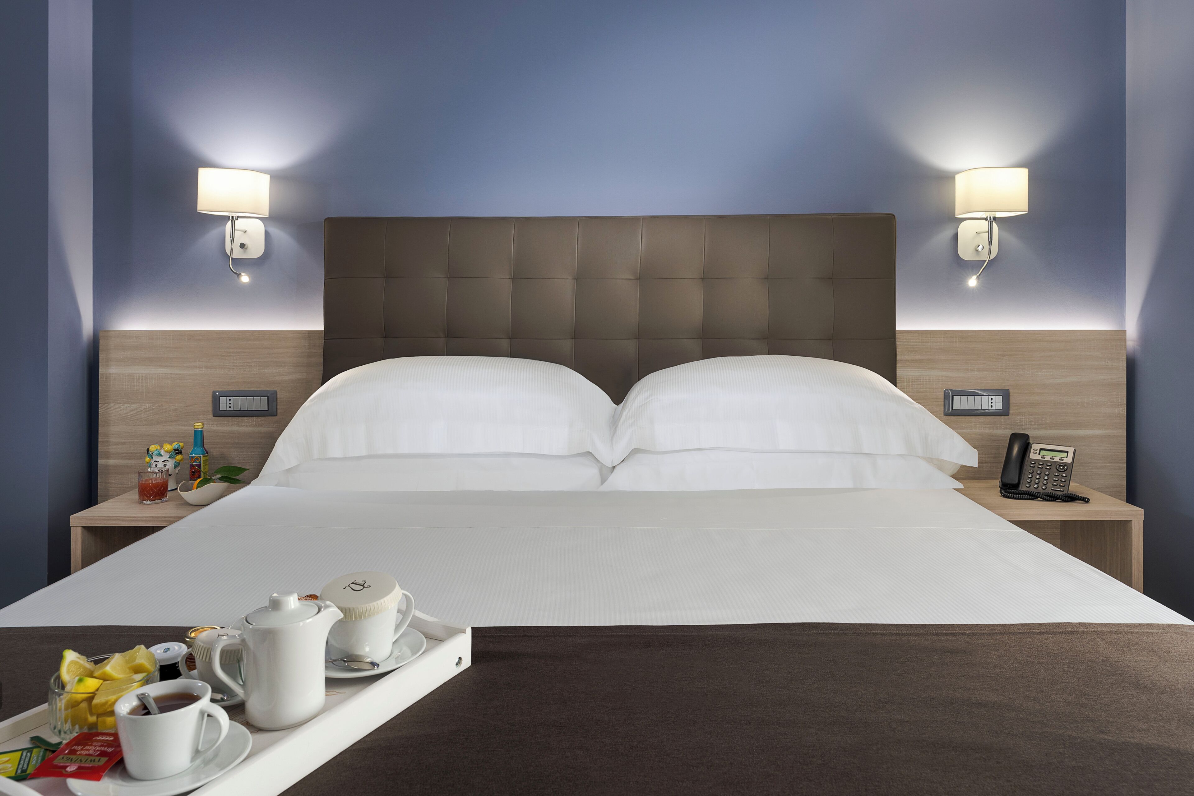 Una cama bien hecha con ropa blanca, un carrito con productos para el desayuno, lámparas de noche y un teléfono en la mesita.