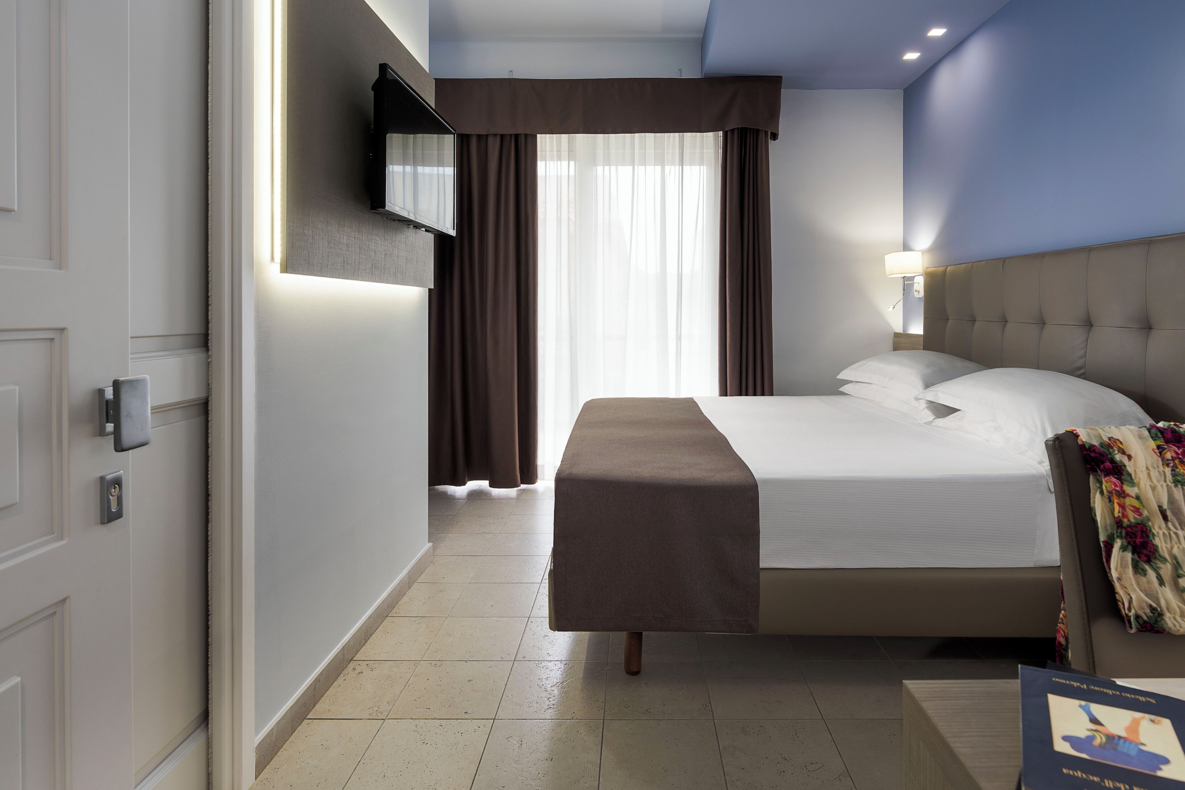 Ein Hotelzimmer mit Bett, Nachttisch, flachbildfernseher an der Wand und einem Fenster mit Vorhängen.