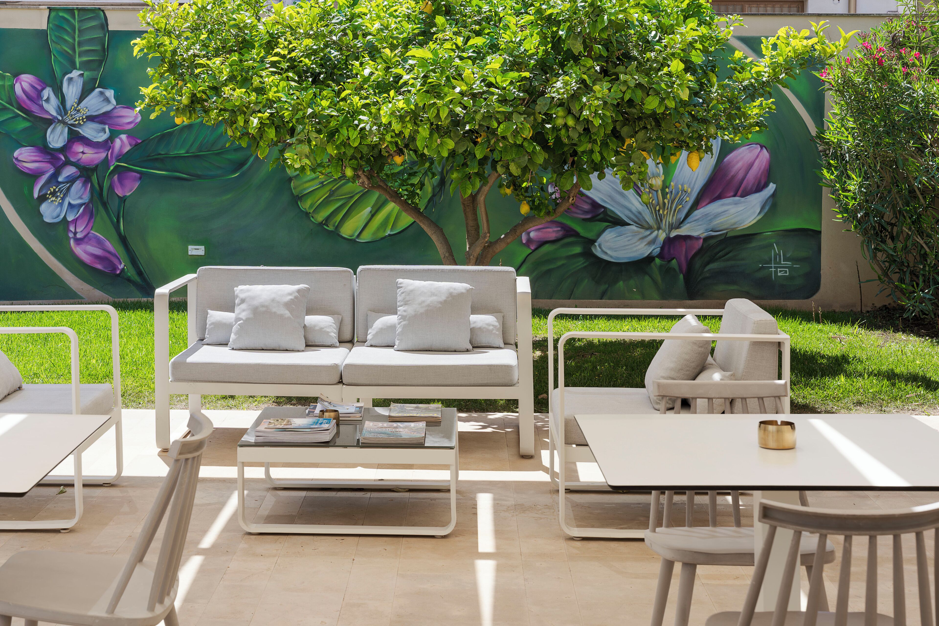 An outdoor seating area with a white sofa and chairs, a small table, and a mural of flowers on the wall.