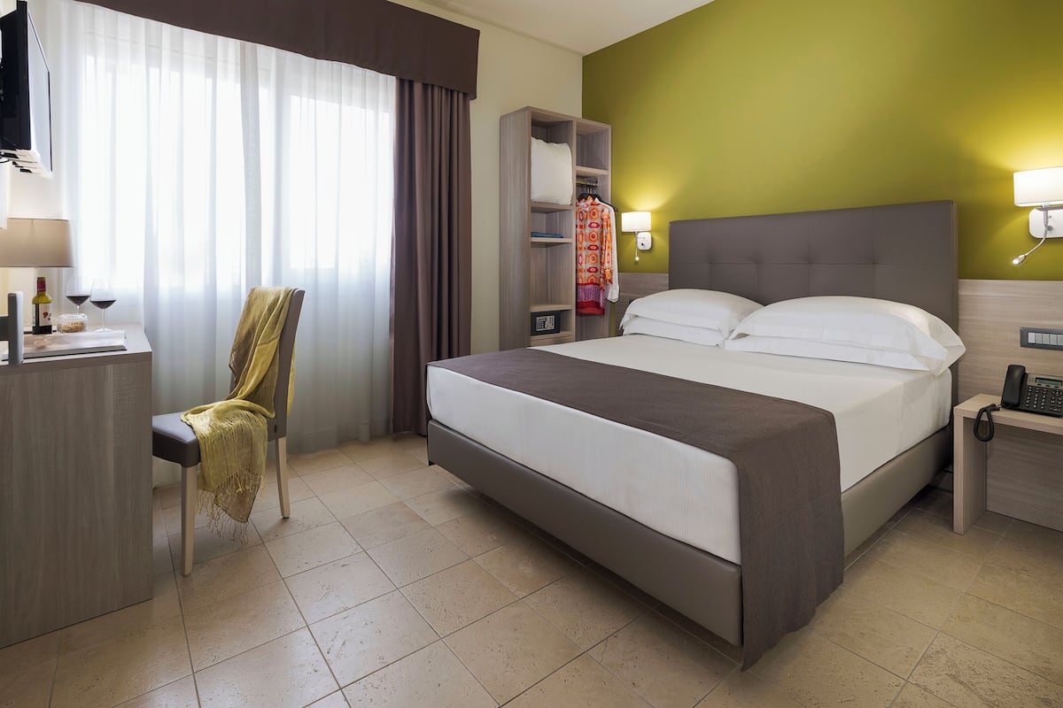 A hotel room with a bed, a chair, a desk with a lamp, a telephone, and a wall-mounted TV.