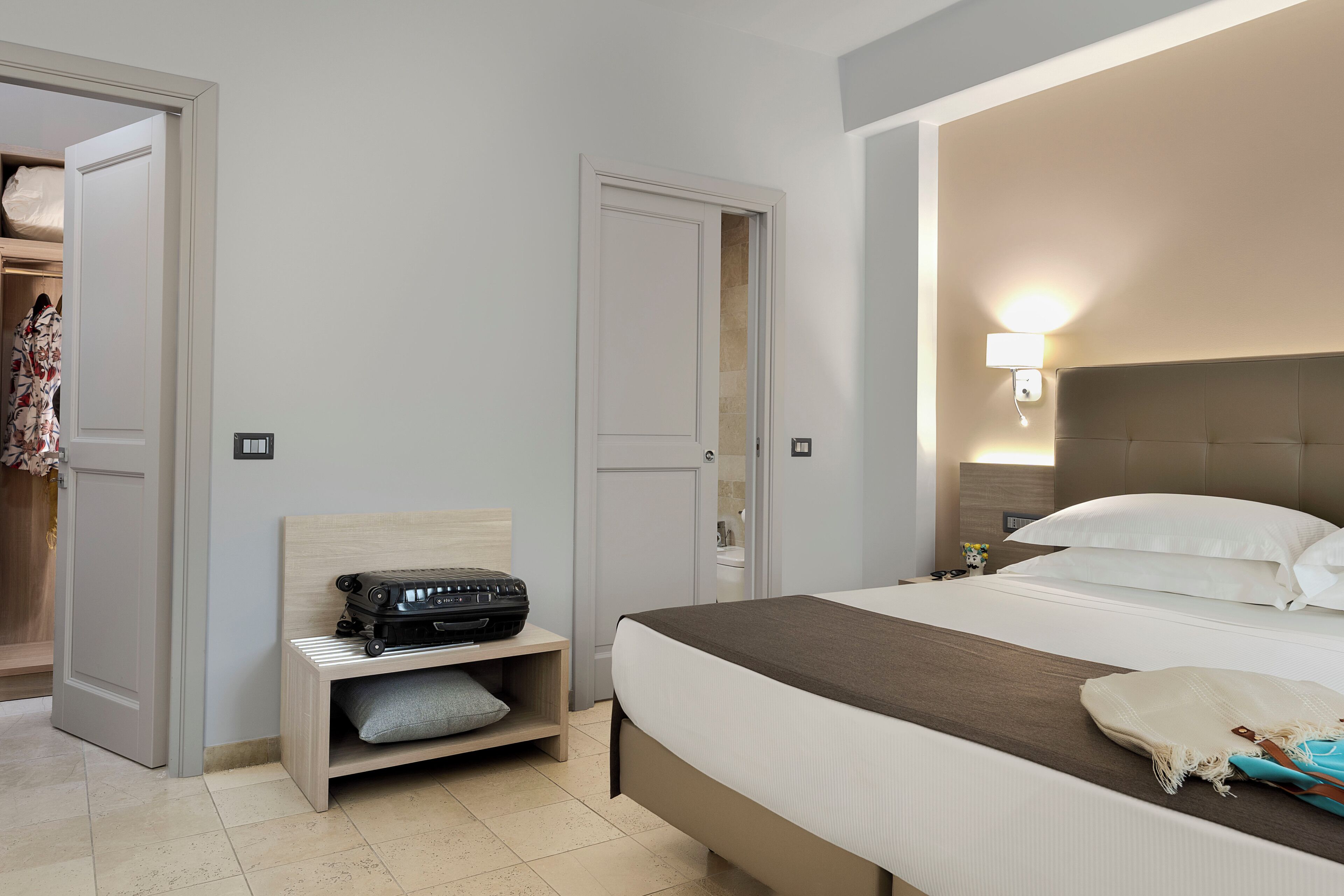 A hotel room with a bed, bedside table, a suitcase, a wardrobe, and a wall-mounted light.