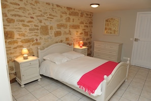 Un dormitorio con pared de piedra, una cama con ropa de cama blanca y una colcha roja, una mesita de noche con una lámpara y un aparador.