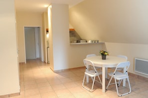 Comedor con una mesa y sillas blancas, una planta en maceta y una cocina al fondo.