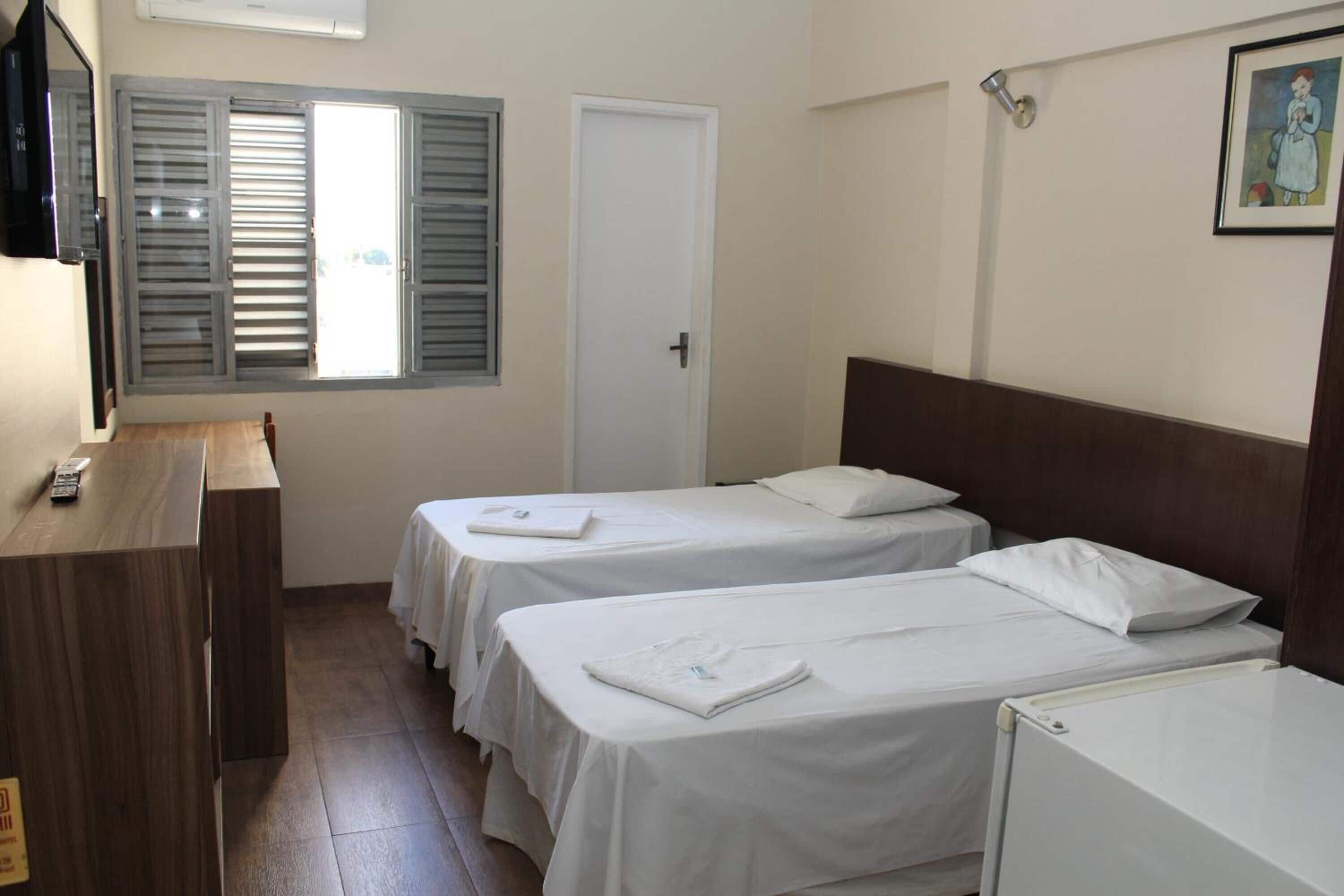A hotel room with two single beds, a wooden desk, a white door, and a window with shutters.