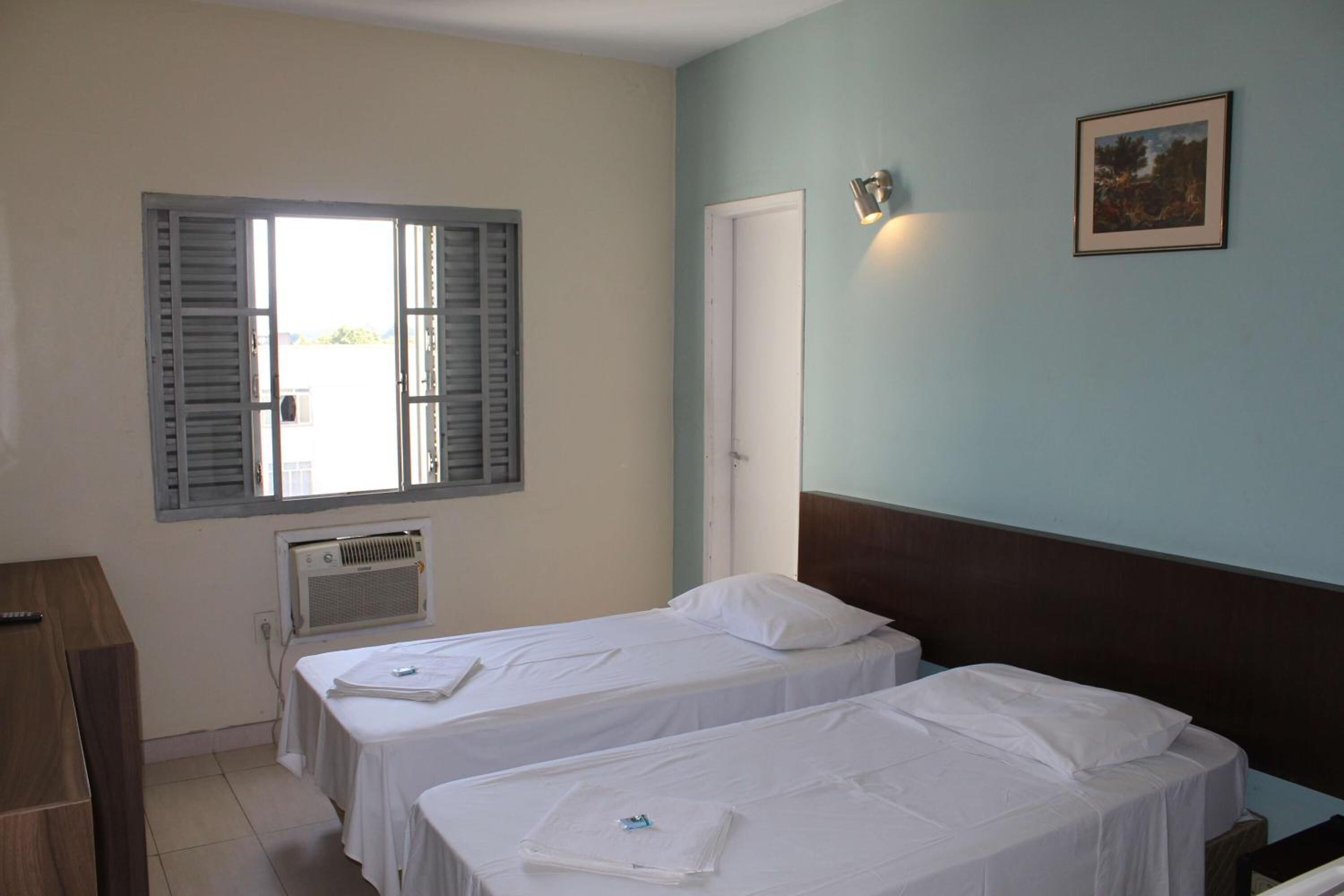 A room with two single beds, a wooden desk, a window with shutters, an air conditioning unit, and a framed picture on the wall.
