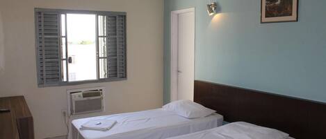A room with two single beds, a wooden desk, a window with shutters, an air conditioning unit, and a framed picture on the wall.