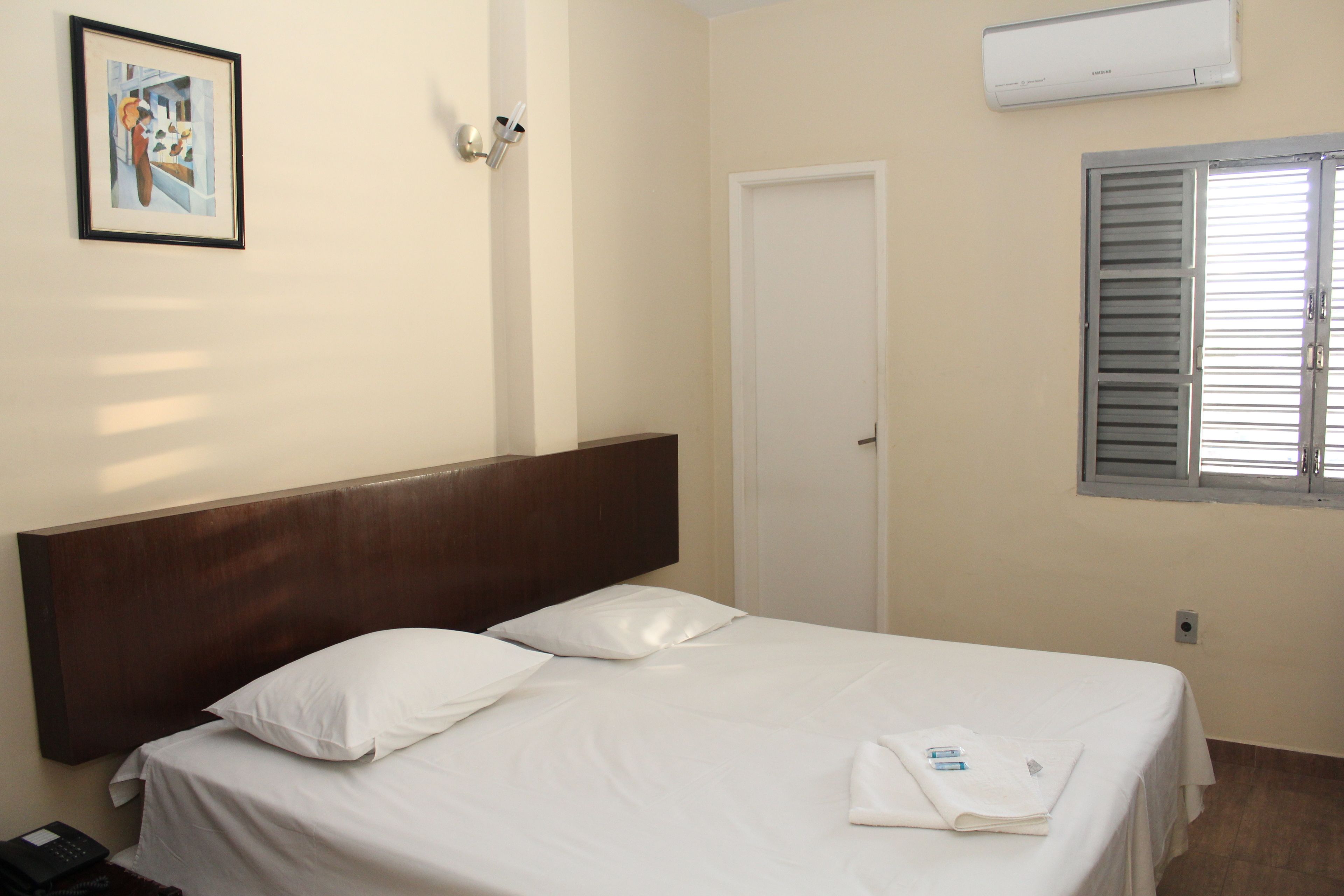 A hotel room with a single bed, white linens, a wooden headboard, a framed picture on the wall, an air conditioning unit, and a window with blinds.