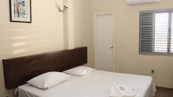 A hotel room with a single bed, white linens, a wooden headboard, a framed picture on the wall, an air conditioning unit, and a window with blinds.