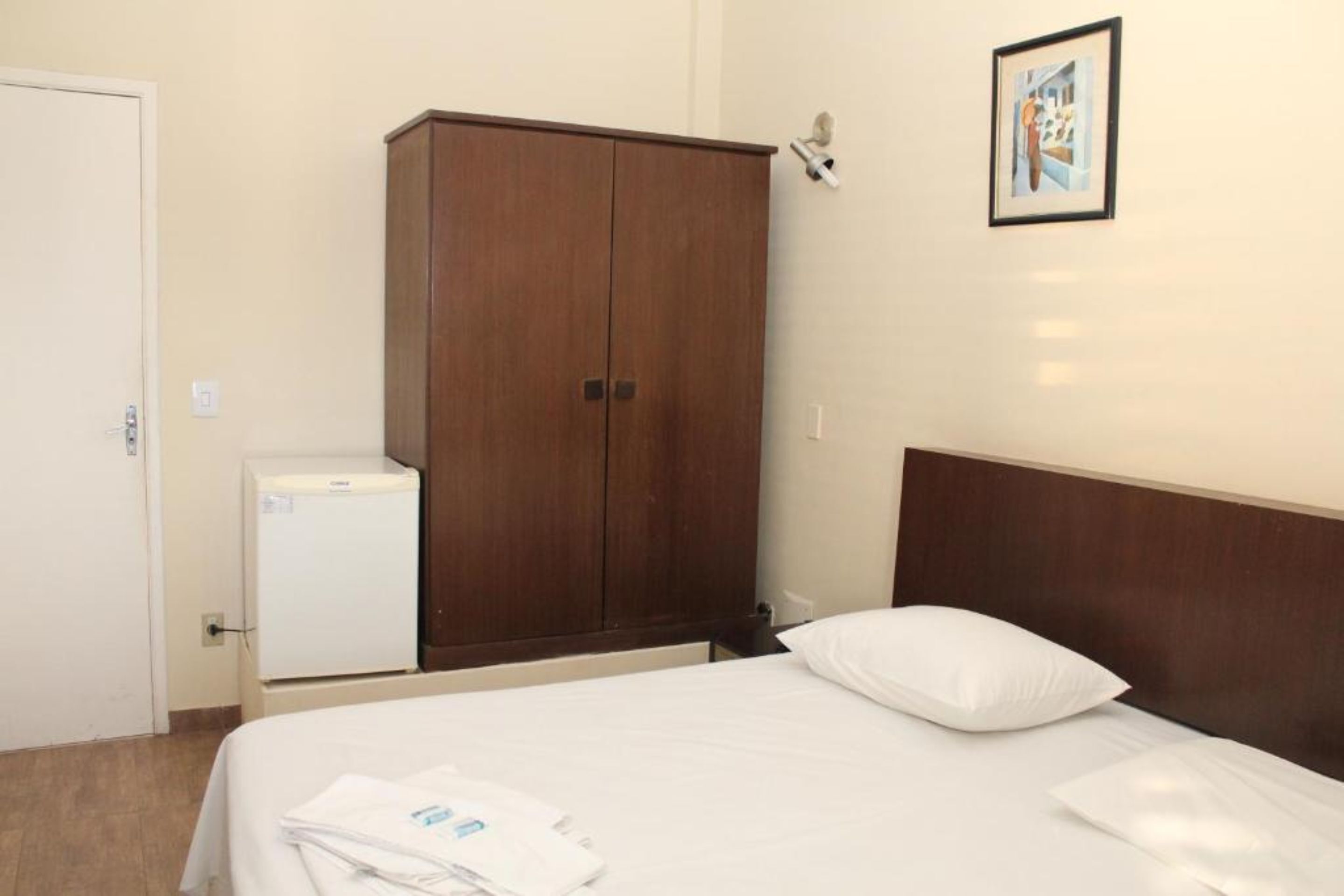 A hotel room with a bed, a wardrobe, a small fridge, and a wall-mounted light fixture.