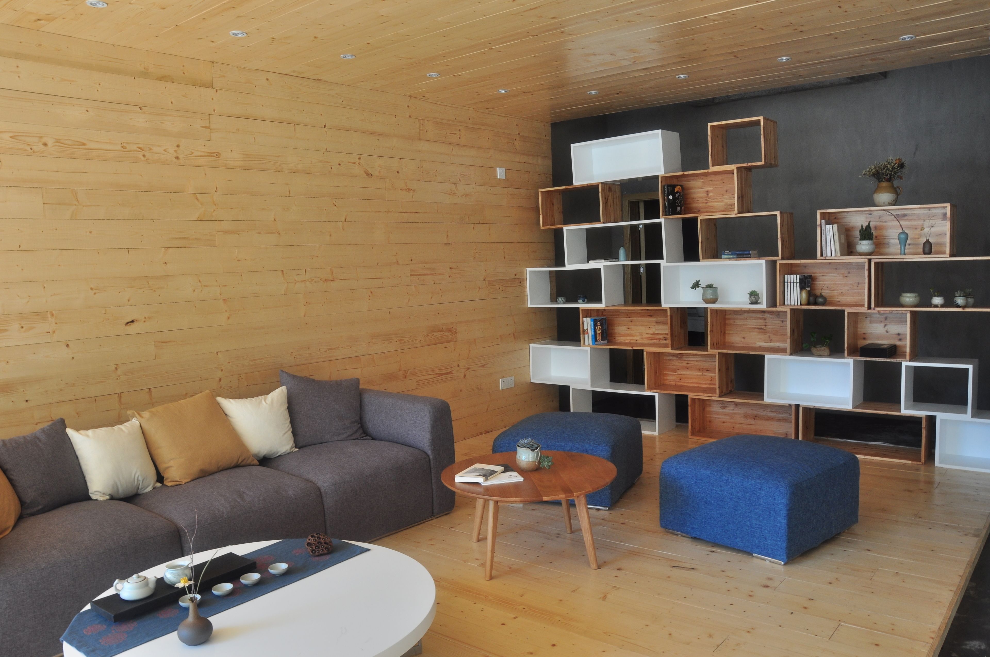 A modern living room with a wooden floor, a grey sofa, a blue ottoman, and a white coffee table. There are wooden shelves with various items on them.