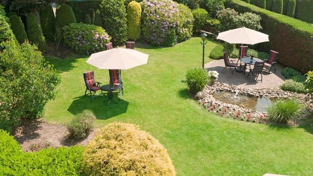 A well-maintained garden with a patio area featuring two tables with chairs and umbrellas, surrounded by neatly trimmed hedges and flowering plants.