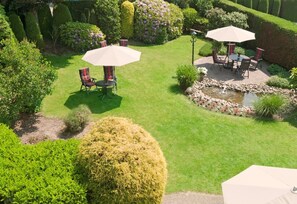 Ein gepflegter Garten mit einer Terrassenanlage, die zwei Tische mit Stühlen und Sonnenschirmen umfasst, umgeben von ordentlich geschnittenen Hecken und blühenden Pflanzen.