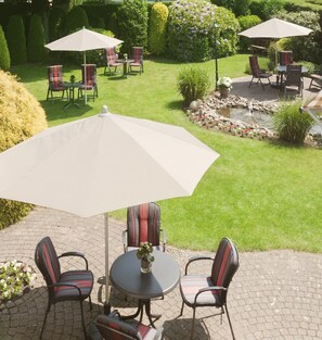 An outdoor patio with a round table and four chairs, each with a striped cushion. There are two large umbrellas providing shade. The patio is paved with bricks and surrounded by well-manicured grass and hedges. - Appartementen-Hotel Geuldal (Epen)