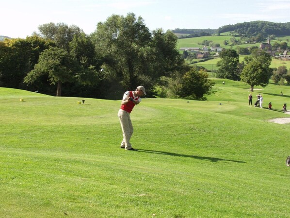 Un campo de golf con un jugador en pleno balanceo, árboles y un pueblo distante.