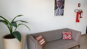 A beige sofa with a patterned pillow, a large potted plant, and a framed portrait on the wall. - Hotel Jeremias (Barretos)