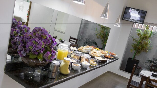 A breakfast buffet with various food items, a vase of purple flowers, and a TV mounted on the wall. - Hotel Jeremias (Barretos)