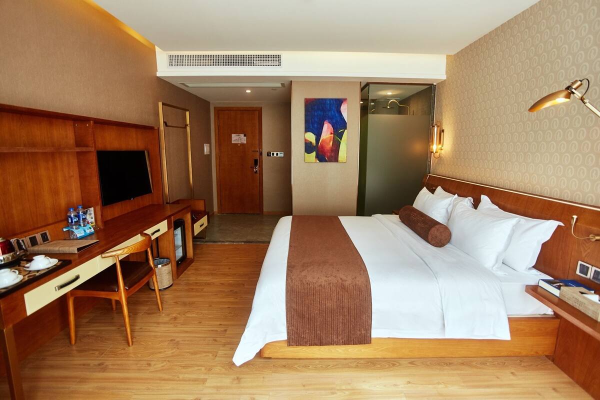 A hotel room with a wooden desk, a bed with white linens, a brown throw blanket, a TV, and a lamp.