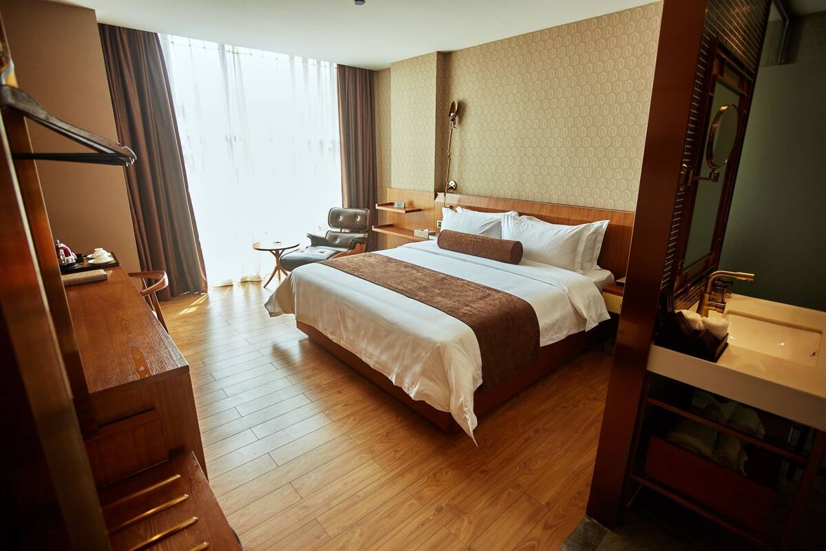 A hotel room with a large bed, wooden floor, a desk, a chair, a nightstand, and a bathroom with a sink and mirror.