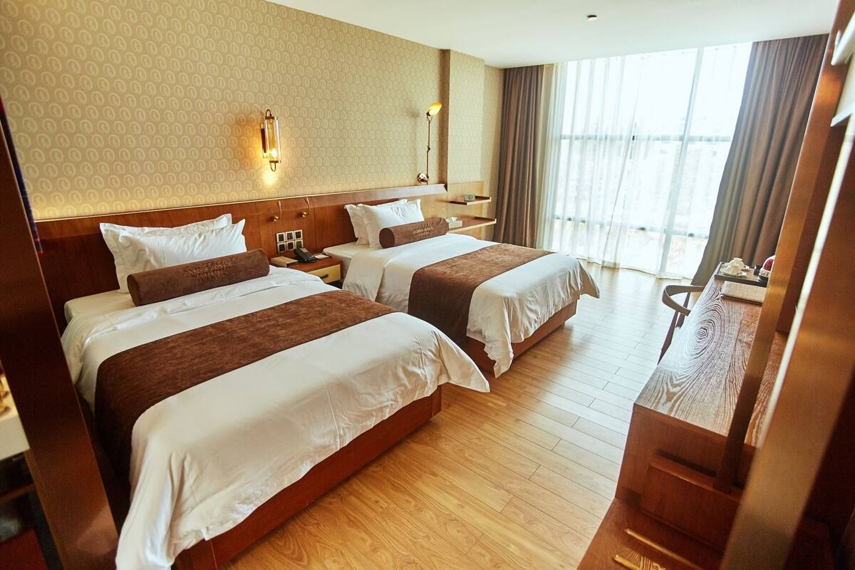 A hotel room with two beds, wooden flooring, a large window with curtains, and a desk with a lamp.