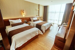 Habitación de hotel con dos camas, suelo de madera, un ventanal grande con cortinas y un escritorio con lámpara.