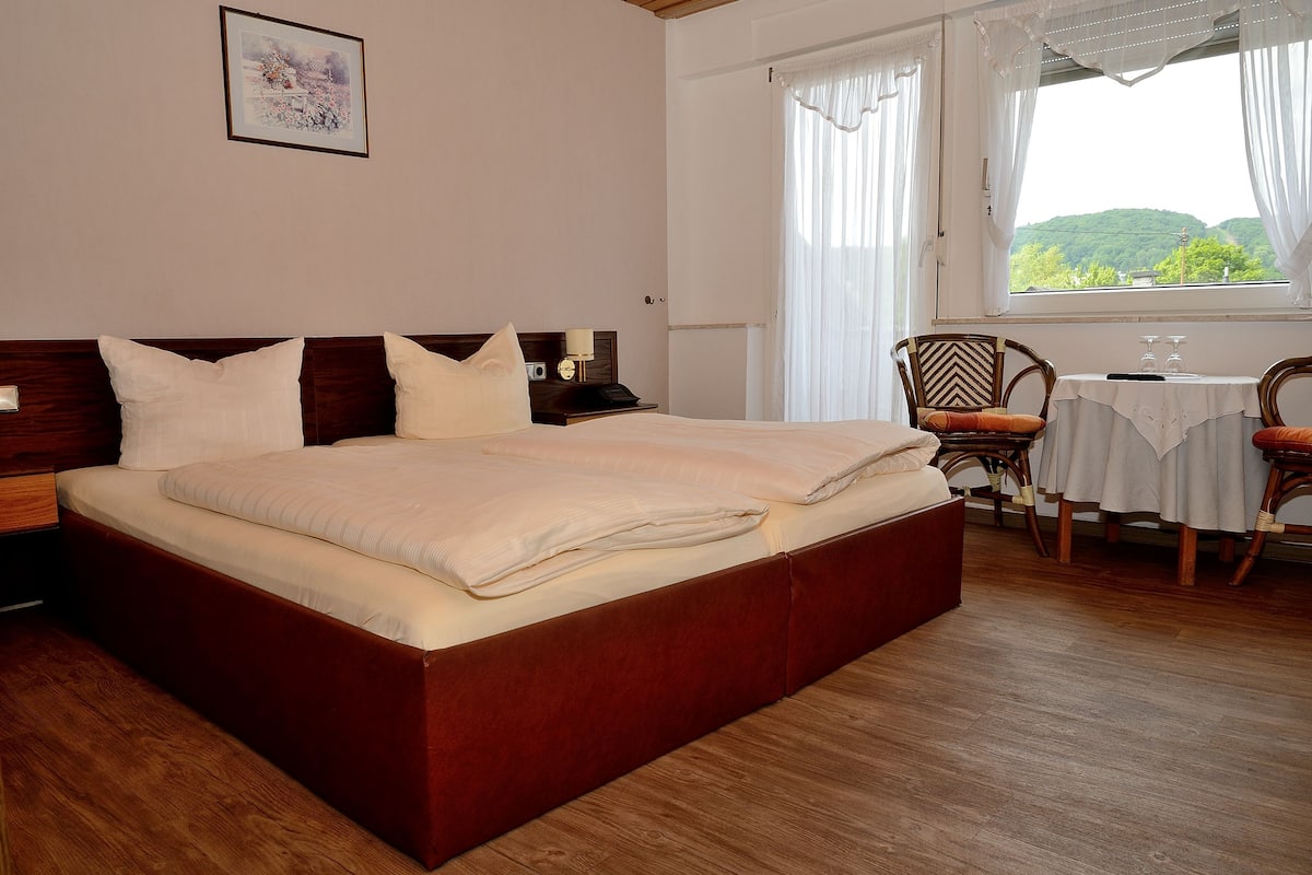 A neatly made bed with a wooden headboard, a bedside table with a lamp, and a chair with a table beside a window with curtains.