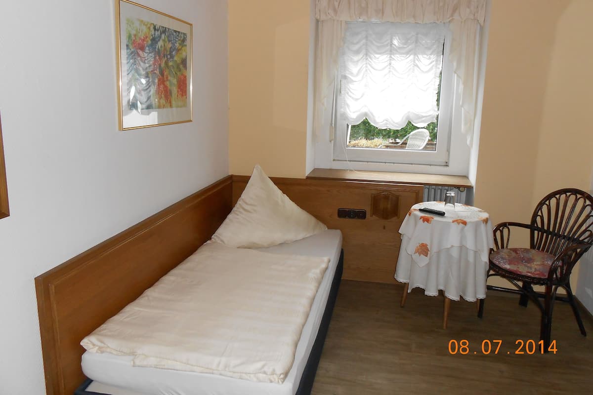 A small, single-bed room with a wooden headboard, a table with a white tablecloth, a chair, and a window with white curtains.