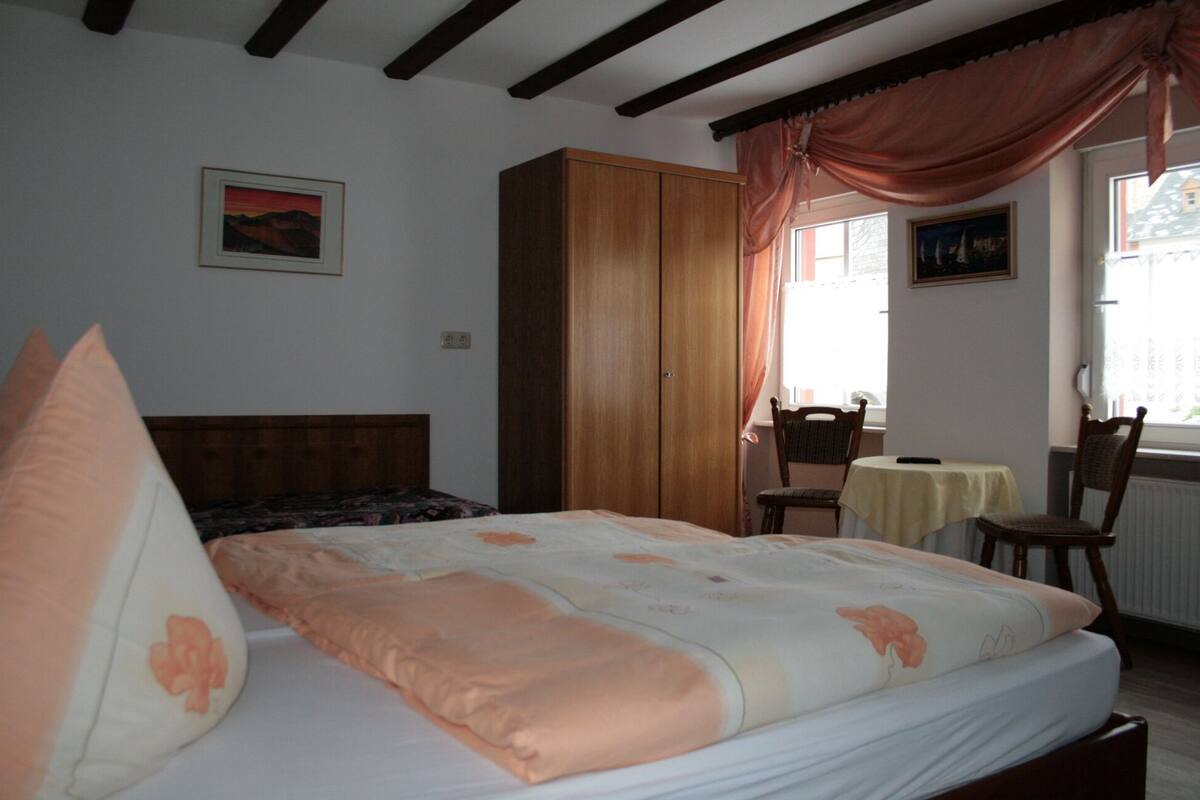 A bedroom with a bed, a wardrobe, a chair, a table, and a window with curtains.