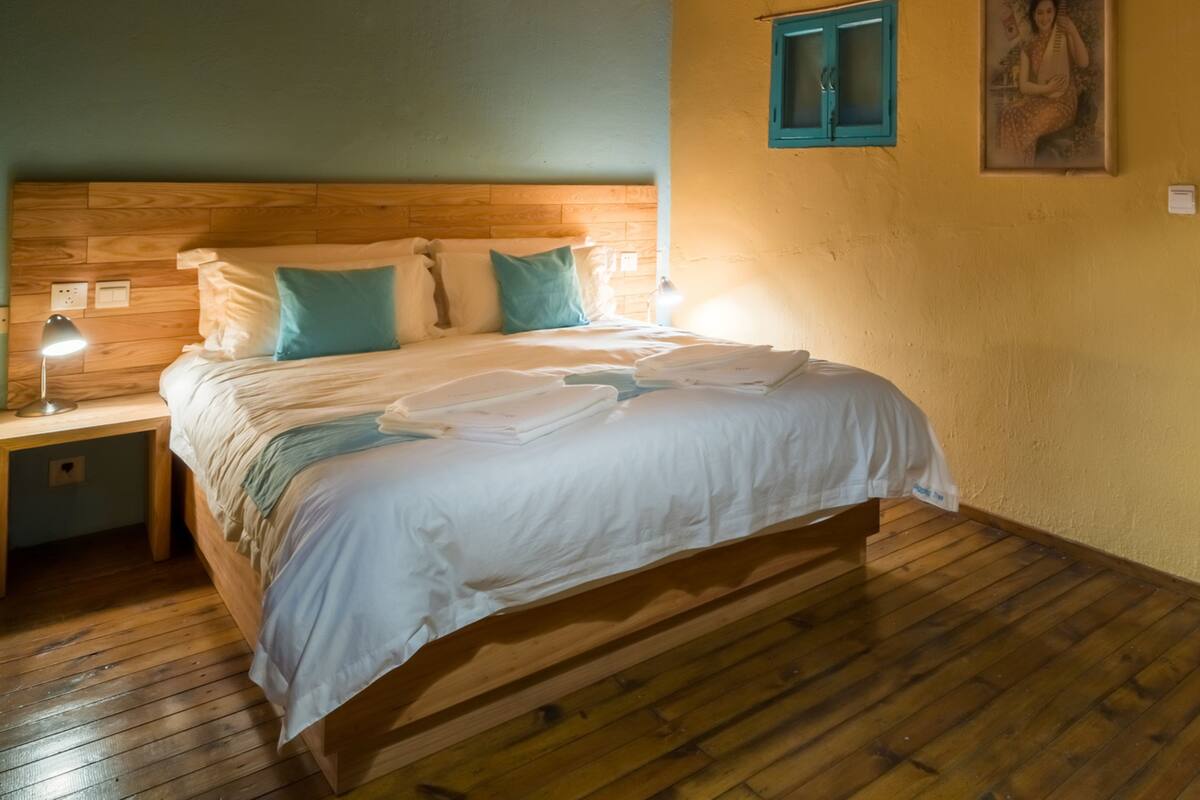A wooden-framed bed with white bedding, a nightstand with a lamp, and a wall-mounted window with blue shutters.