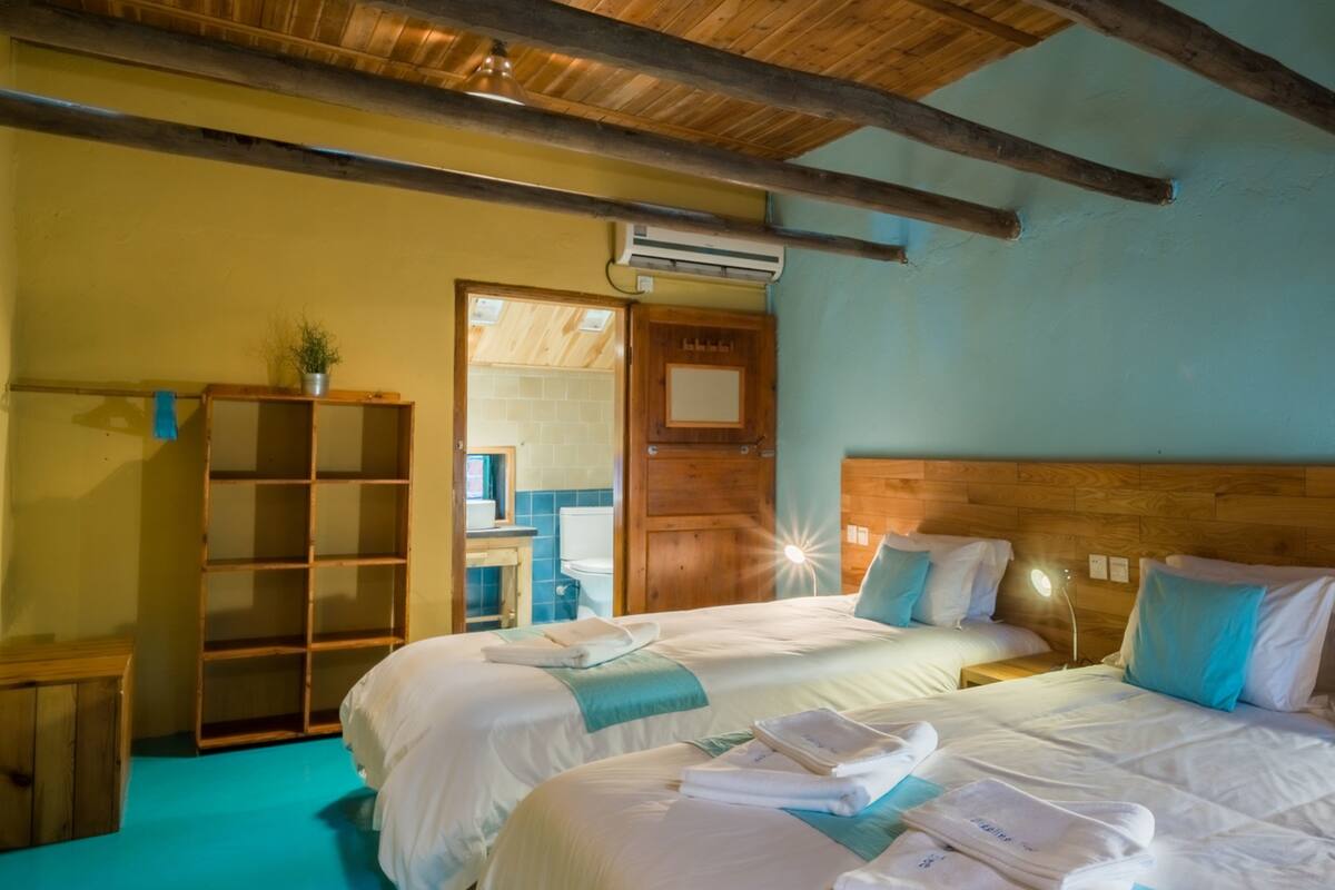 A room with two beds, wooden beams on the ceiling, a wooden wardrobe, and a bathroom visible through an open door.