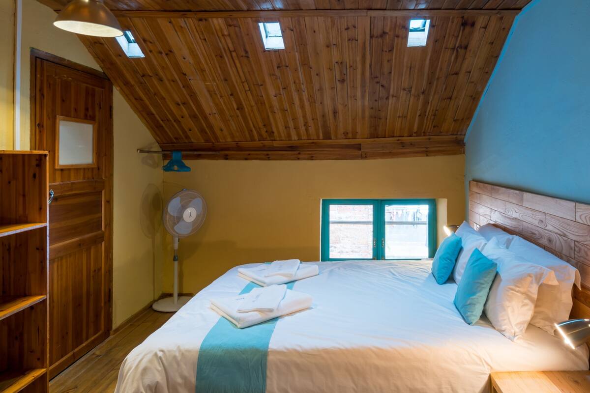 A bedroom with a wooden ceiling, a bed with white bedding and blue pillows, a standing fan, and a window with a view of the outside.
