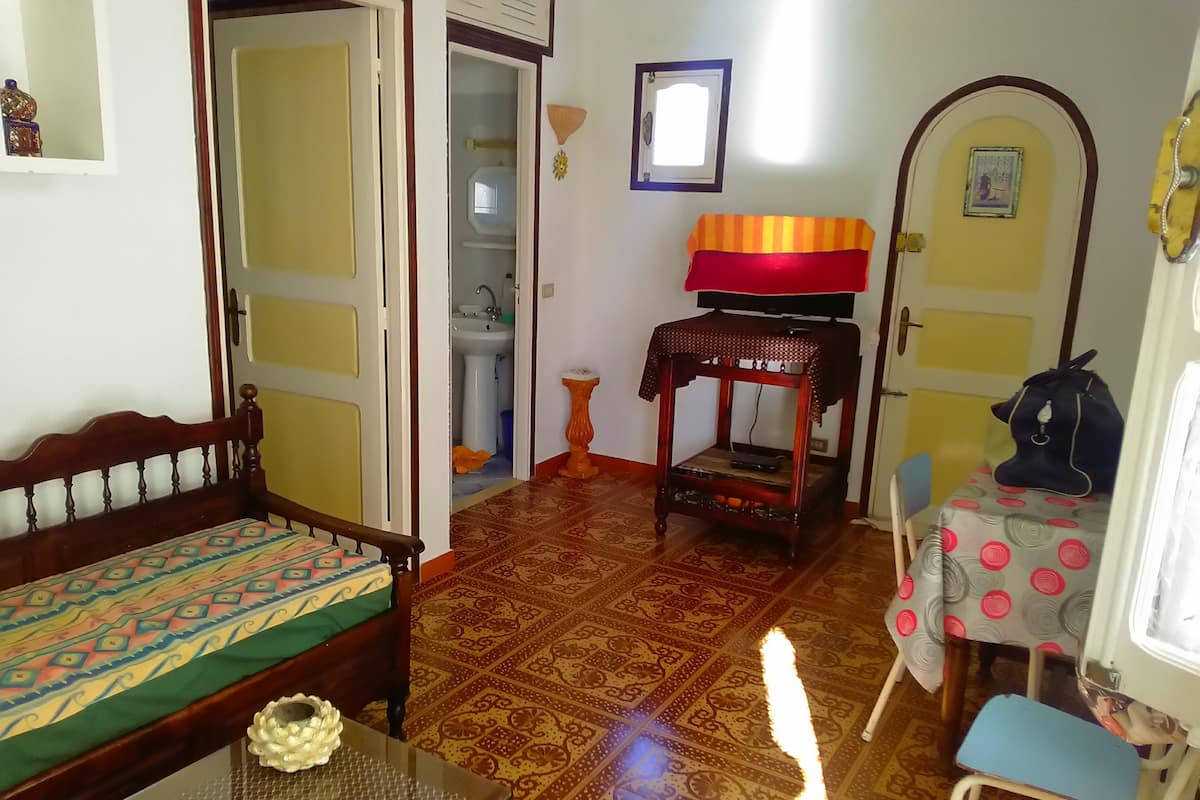 Une pièce comprenant un lit, une petite table ornée d’un objet décoratif, une chaise, une table d’appoint en bois avec une lampe, et un téléviseur posé sur un meuble.