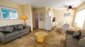 Uma sala de estar com sofá, mesa de centro, área de jantar com cadeiras, geladeira e ventilador de teto.