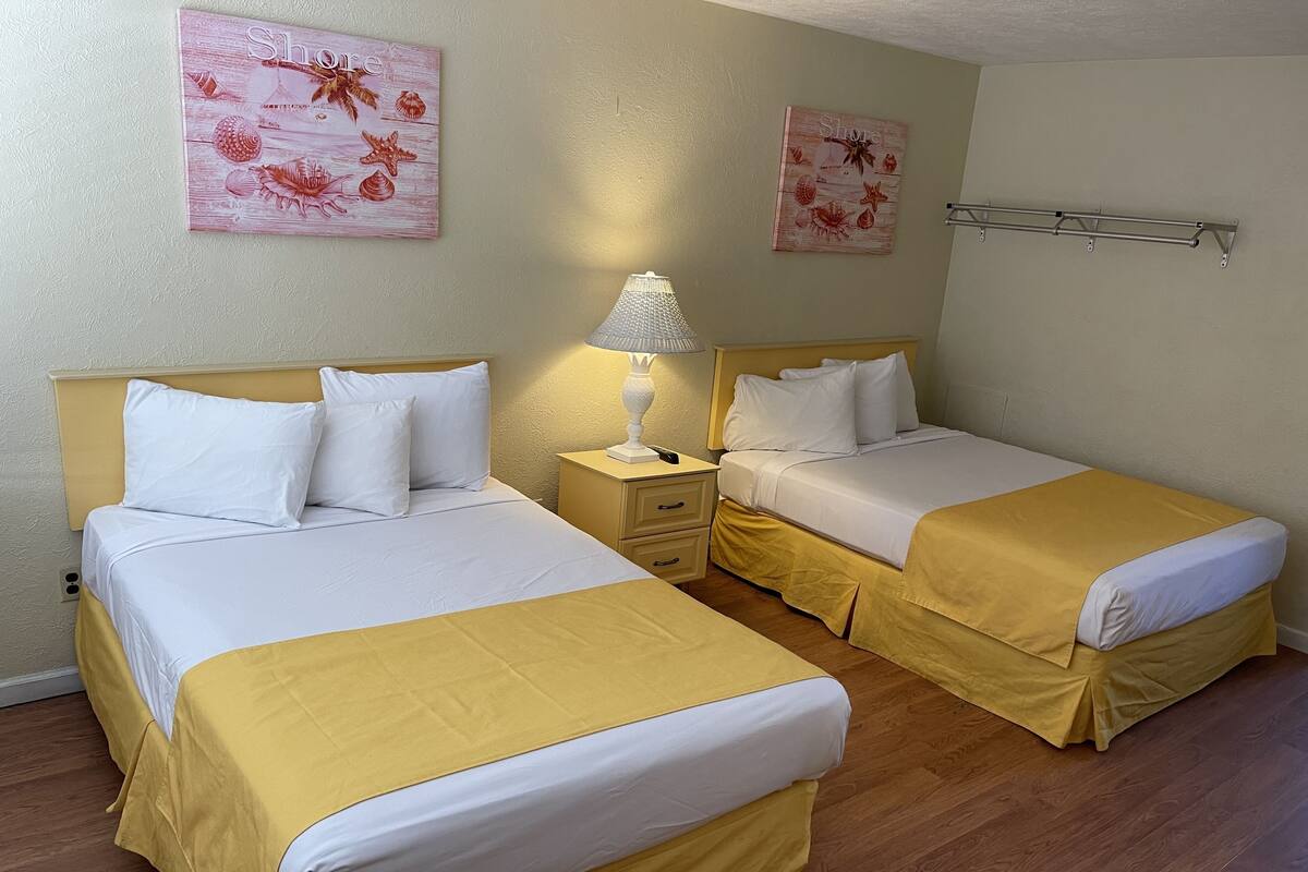 A hotel room with two beds, each with a yellow bedspread and white linens. There are two bedside tables with lamps, and the walls are adorned with floral artwork.