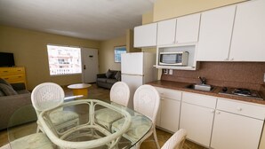 Uma cozinha compacta com armários brancos, micro-ondas e pia. Há uma área de refeições com mesa de vidro e cadeiras de vime.